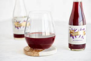 The Saskatoon, a dark red drink, in a stemless wine glass, with a bottle beside it