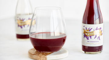 The Saskatoon, a dark red drink, in a stemless wine glass, with a bottle beside it