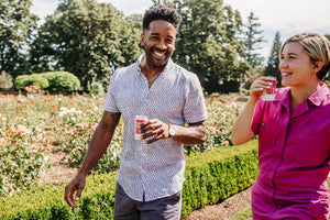 A man and a woman walk through a rose garden on a sunny day, smiling and holding a canned non-alcoholic cocktail.