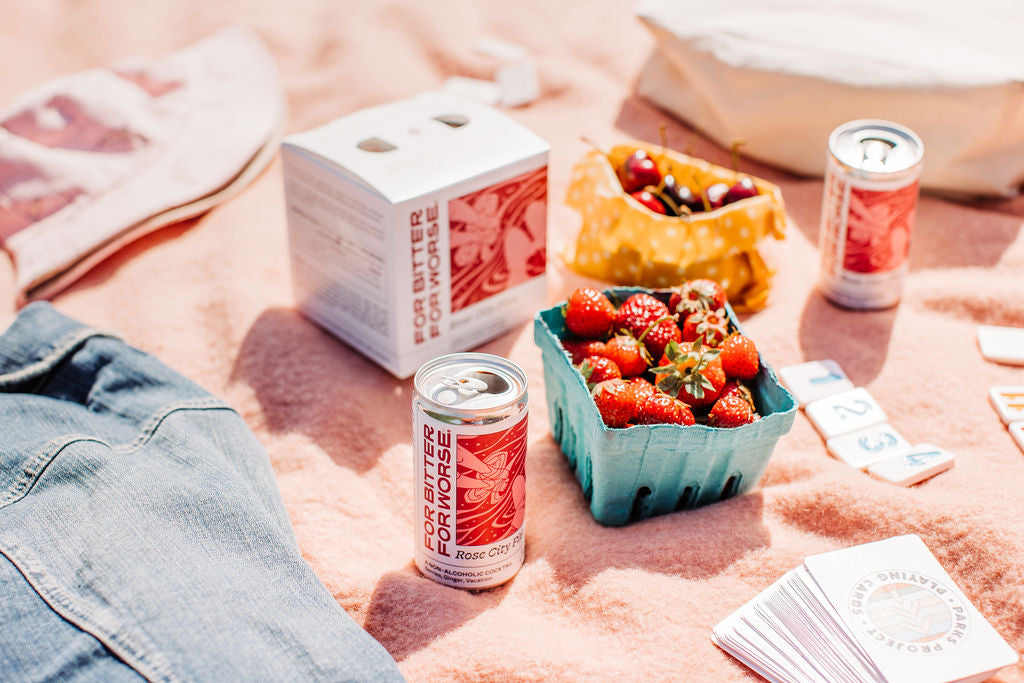 Arranged on a pale pink picnic blanket are a basket of strawberries, two cans containing a non-alcoholic cocktail, a bag of cherries, and a deck of playing cards.