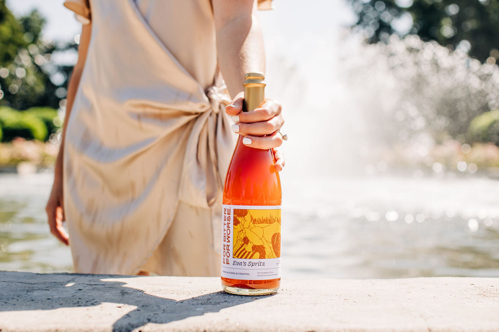 A woman holds a bottle of Eva's Spritz, a non-alcoholic cocktail from For Bitter For Worse, in front of a fountain.