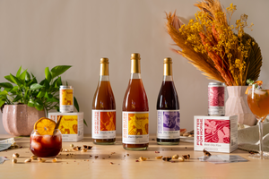 Three bottled non-alcoholic cocktails and two four-packs of cans with variably colored labels stand on a wooden table.