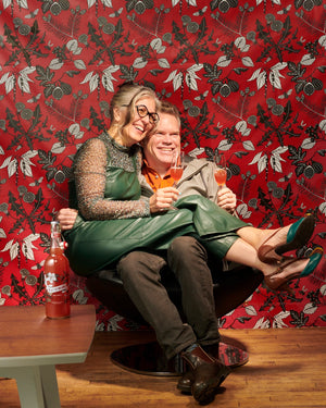 A smiling man wearing an orange dress shirt sits in a chair. A smiling woman wearing a green dress sits across his lap. Behind them is red wallpaper with black-and-white cut paper art depicting various botanicals.