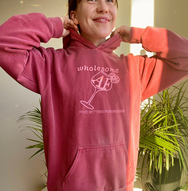 A young woman wearing a dark dusty rose colored sweatshirt. The sweatshirt depicts a wine glass with a citrus wheel garnish and reads "wholesome AF."