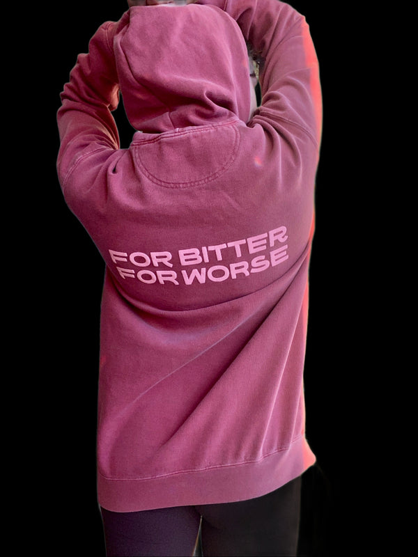 A young woman wearing a dark dusty rose colored sweatshirt. The back of the sweatshirt reads "For Bitter For Worse."