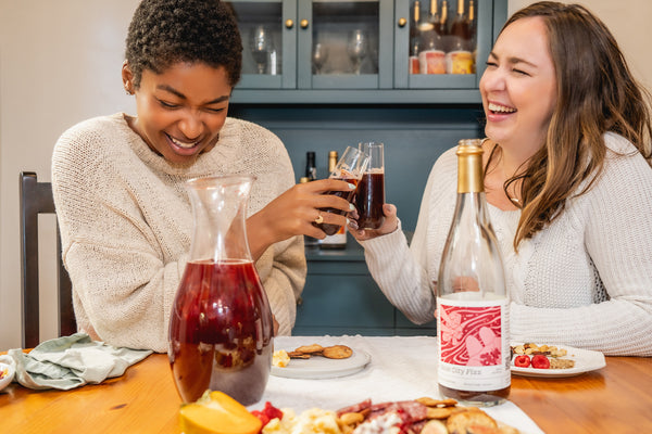 Two laughing women sit at a table, clinking together glasses of non-alcoholic mixed drinks. A charcuterie board and a carafe containing a non-alcoholic cocktail rest on the table.