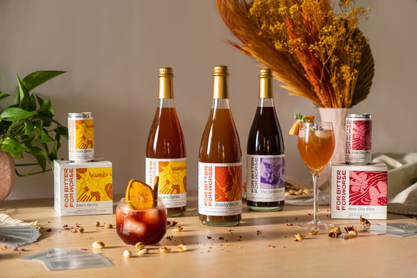 Three bottled non-alcoholic cocktails and two four-packs of cans with variably colored labels stand on a wooden table.