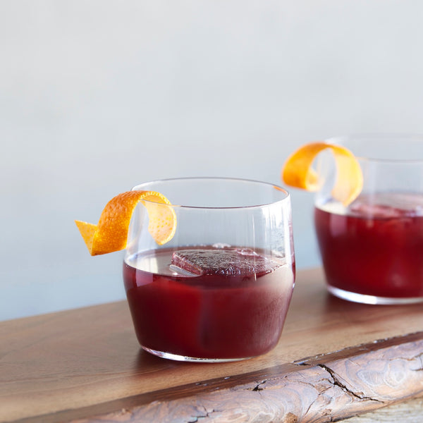 Close-up of two short glasses of a cherry-red non-alcoholic cocktail with orange peel garnishes.