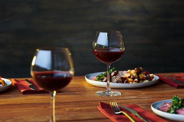 Close-up of two wine glasses containing a non-alcoholic cocktail that rest on a wood dining table next to plates of food.