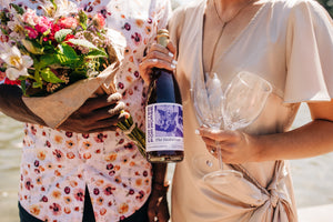 Close up of the torsos of a man and a woman. The man holds a bouquet and the woman holds two wine glasses in one hand and a bottle containing a non-alcoholic beverage in the other.