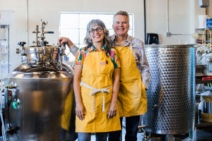 Shelley and Jeff, the founders of For Bitter For Worse, wear bright yellow aprons and smile as the stand between two large stainless steel mixing tanks.