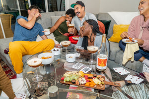 Five people laugh as they sit on a couch around a glass coffee table that holds a charcuterie board, mocktails in martini glasses, playing cards, and a bottle containing a non-alcoholic beverage.