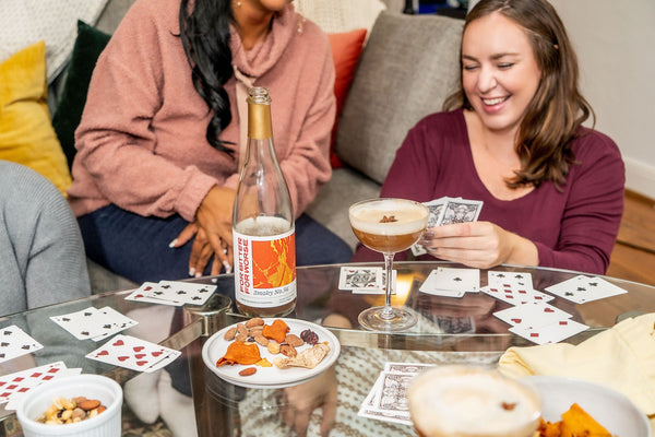Two women play cards at a coffee table upon which are snacks and a non-alcoholic beverage