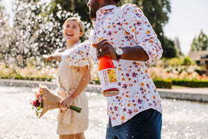 A man and a woman laugh as they wade through a fountain. The woman holds a bouquet and the man holds a bottle of Eva's Spritz.